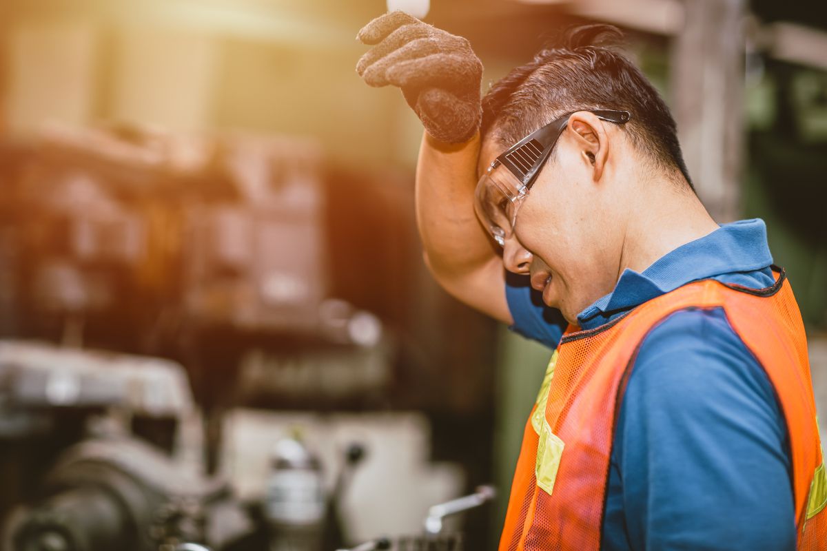 Man in orange vest and safety glasses wiping his head with his forearm